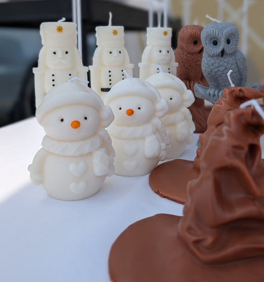 Assorted candles, including snowman figure candle, a hat, an owl, with nutcrackers behind on a white surface.